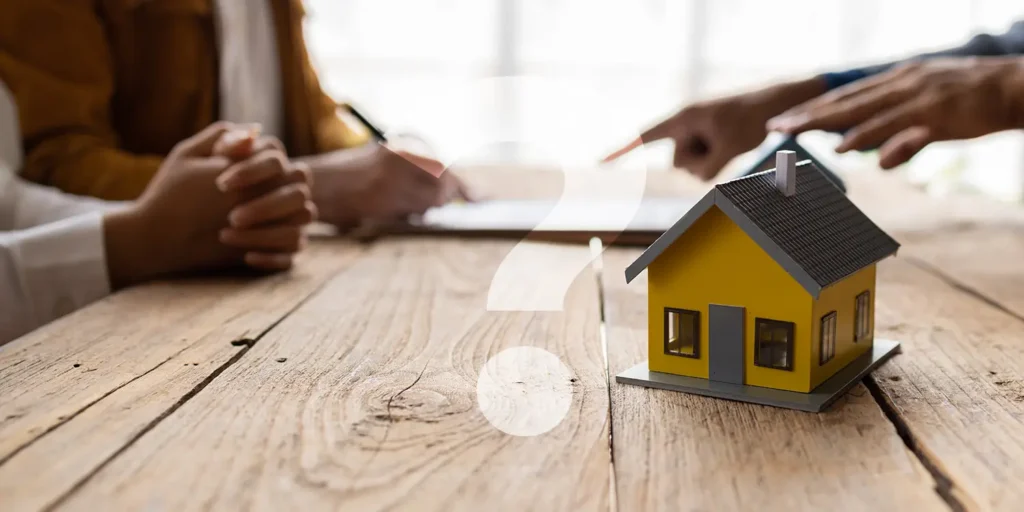 Conveyancing FAQs. Model house on wooden desk with people discussing conveyancing in the background