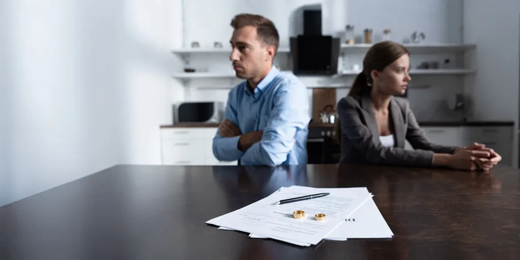 A separated couple sitting back-to-back at a table with divorce papers and wedding rings in the foreground, symbolising financial settlement and legal separation.