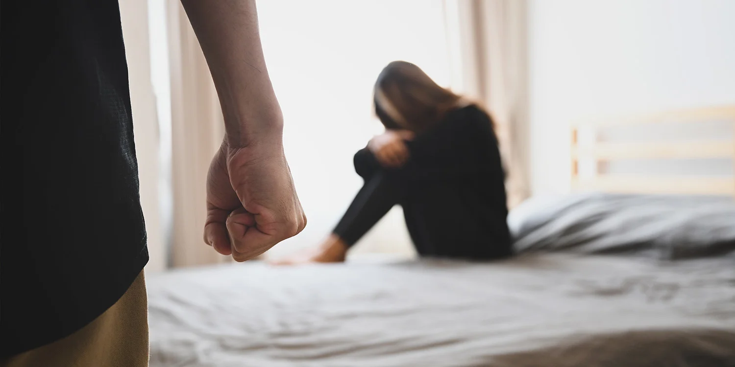 Man with clenched fist in foreground and distressed woman on bed, representing domestic abuse in family proceedings
